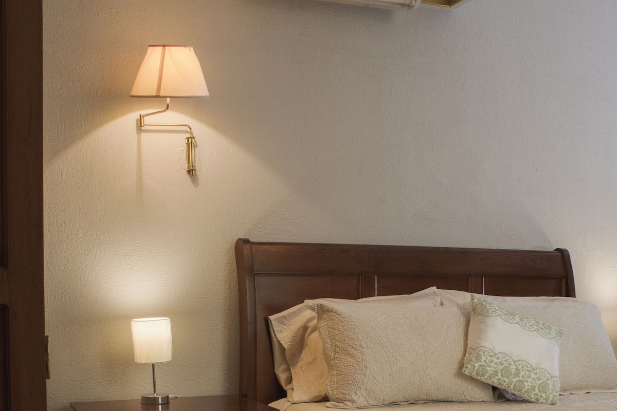 A neatly made bed with a wooden headboard, a bedside table with a lamp, and a wall-mounted air conditioner.