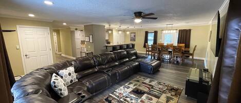 A spacious living room with a large leather sectional sofa, a dining area with wooden chairs, and a kitchen in the background.