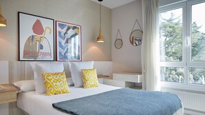 Un dormitorio moderno con una cama grande, dos almohadas, un edredón azul, una mesita de noche de madera, una lámpara colgante y un espejo en la pared, además de una ventana con vistas a árboles.