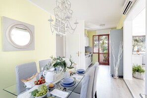 A modern dining area with a glass table set for four, surrounded by grey upholstered chairs. A white chandelier hangs above, and a large window provides natural light.