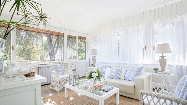 A bright, white room with a glass coffee table, wicker furniture, and large windows with sheer curtains.