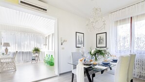 A modern dining area with a glass table, white chairs, and a chandelier. There are framed pictures on the wall and a plant on the table.