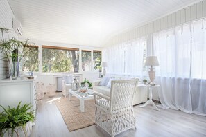 A bright, airy room with white wicker furniture, large windows, and a view of greenery outside.