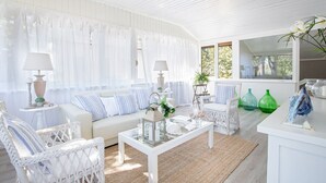A bright, airy room with a white sofa, two white wicker chairs, a glass coffee table, and a small side table with a lamp.