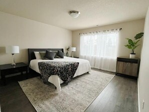 Un dormitorio ordenado con cama, mesitas de noche, una planta en maceta y una ventana con cortinas.