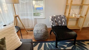A living room with a sofa, ottoman, floor lamp, and a chair with a patterned throw.