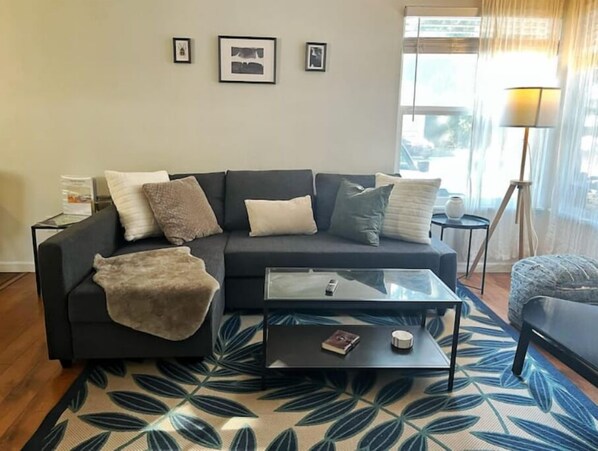 Un salon moderne avec un canapé gris foncé, une table basse en verre et un tapis à motifs. Des tableaux encadrés ornent le mur, et une lampe est posée sur une table d’appoint.