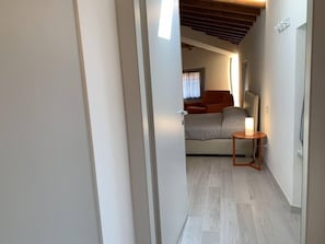 Un couloir étroit mène à une chambre à coucher qui contient un lit, une table de chevet avec une lampe et une fenêtre avec des rideaux.