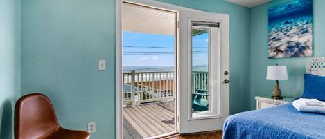 A bedroom with a wooden floor, a bed with a blue bedspread, a brown chair, a small table with a lamp, and a door leading to a balcony with a view of the ocean.