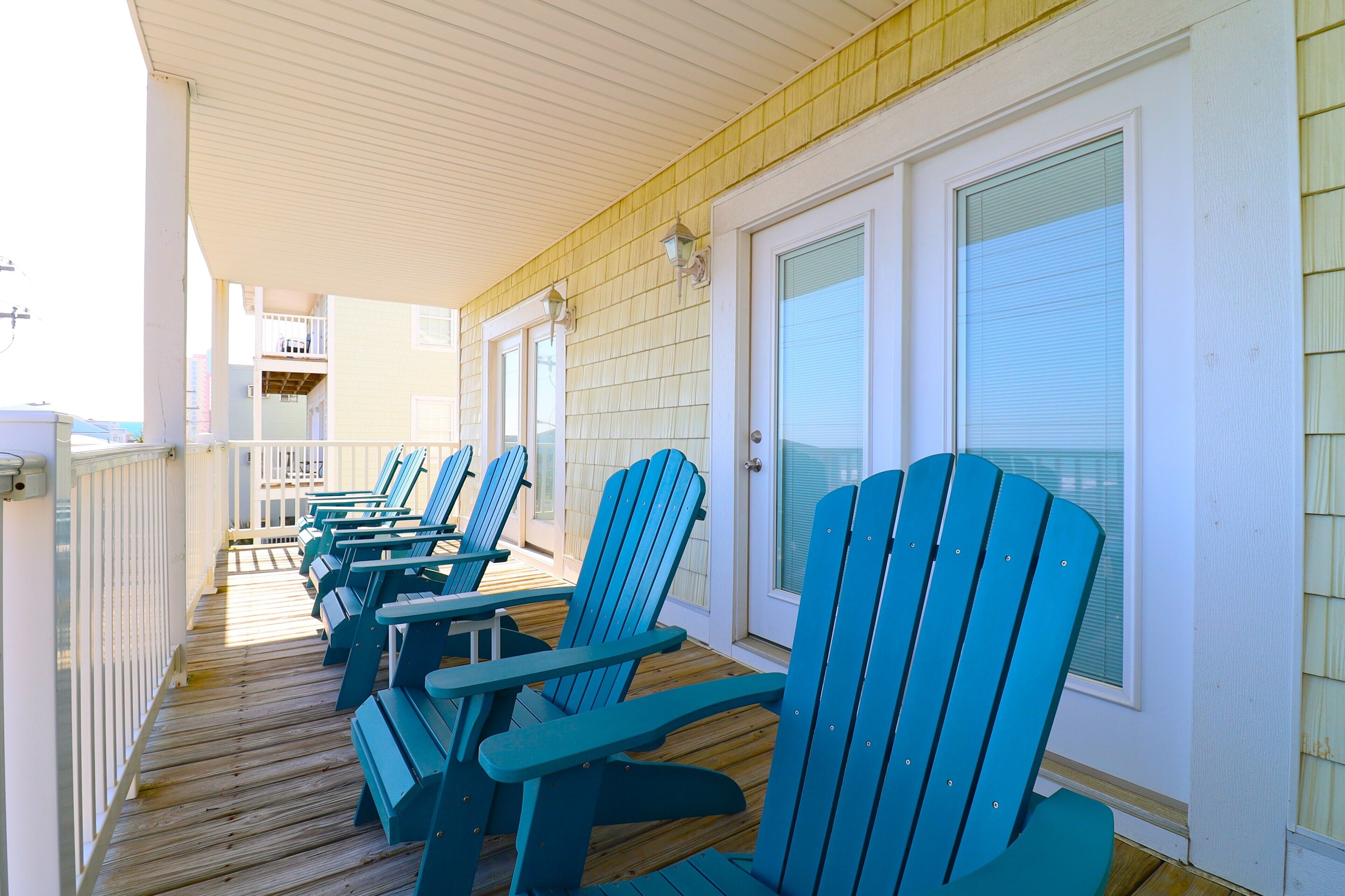 Une terrasse couverte, dotée de plusieurs chaises Adirondack et d’une table, d’une balustrade blanche et d’une porte avec une fenêtre.