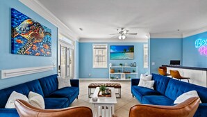 Un salon avec des canapés bleus, une table basse et un téléviseur. On y trouve un ventilateur de plafond, un néon et un tableau sur le thème de la plage accroché au mur.