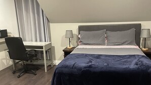A bedroom with a bed, two bedside tables, a desk, a chair, and a window with curtains. - Cozy Downtown Studio (Moncton)
