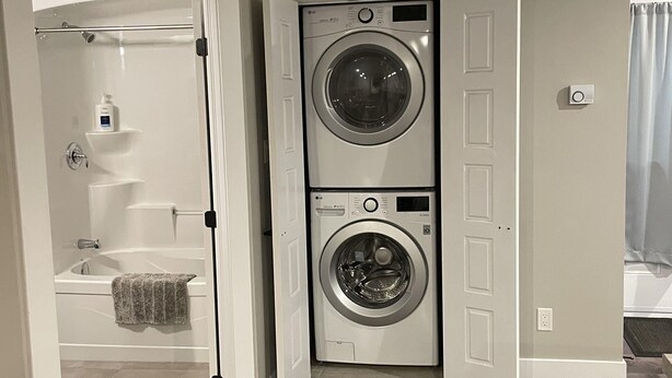 A laundry room with two stacked front-loading washing machines, a door leading to a bathroom, and a visible light switch on the wall. - Cozy Downtown Studio (Moncton)