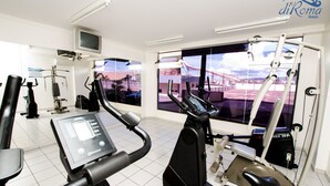 A modern gym with various exercise equipment, a television, and large windows offering views of a bridge and cityscape. - Villas diRoma com um dia no Acqua Park (Caldas Novas)