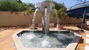 A square water feature with three streams of water flowing from a central stone structure, surrounded by potted plants and a boat. - Villas diRoma com um dia no Acqua Park (Caldas Novas)