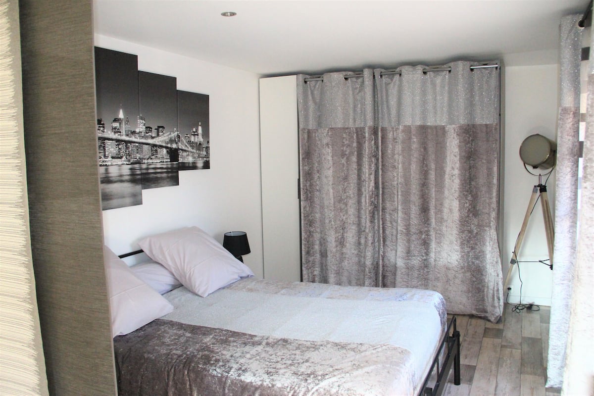 A bedroom with a bed, a nightstand, a lamp, a curtain, and a wall-mounted mirror.