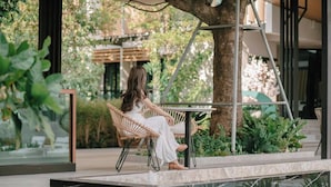 Una mujer sentada en una silla de mimbre junto a una mesa, en un ambiente exterior moderno con un árbol y follaje de fondo.