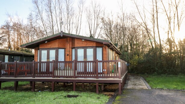A wooden cabin with a balcony, surrounded by trees and a grassy area. - The Goodwood Annex (Chichester)