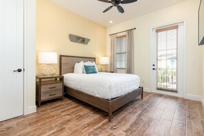 Un dormitorio con una cama de madera, dos mesitas de noche con lámparas, un ventilador de techo, una puerta con ventana y un adorno en la pared.