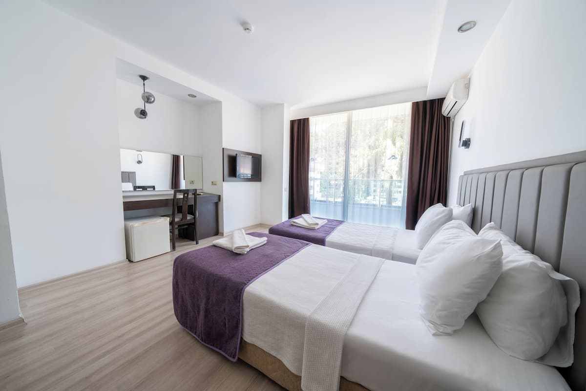 A hotel room with two beds, a desk, a mirror, a TV, and a balcony with curtains.
