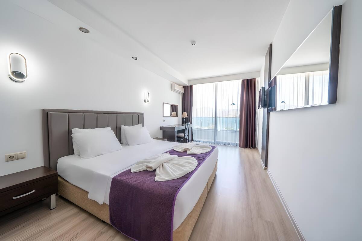 A modern hotel room with a large bed, a desk, a chair, and a balcony with curtains.