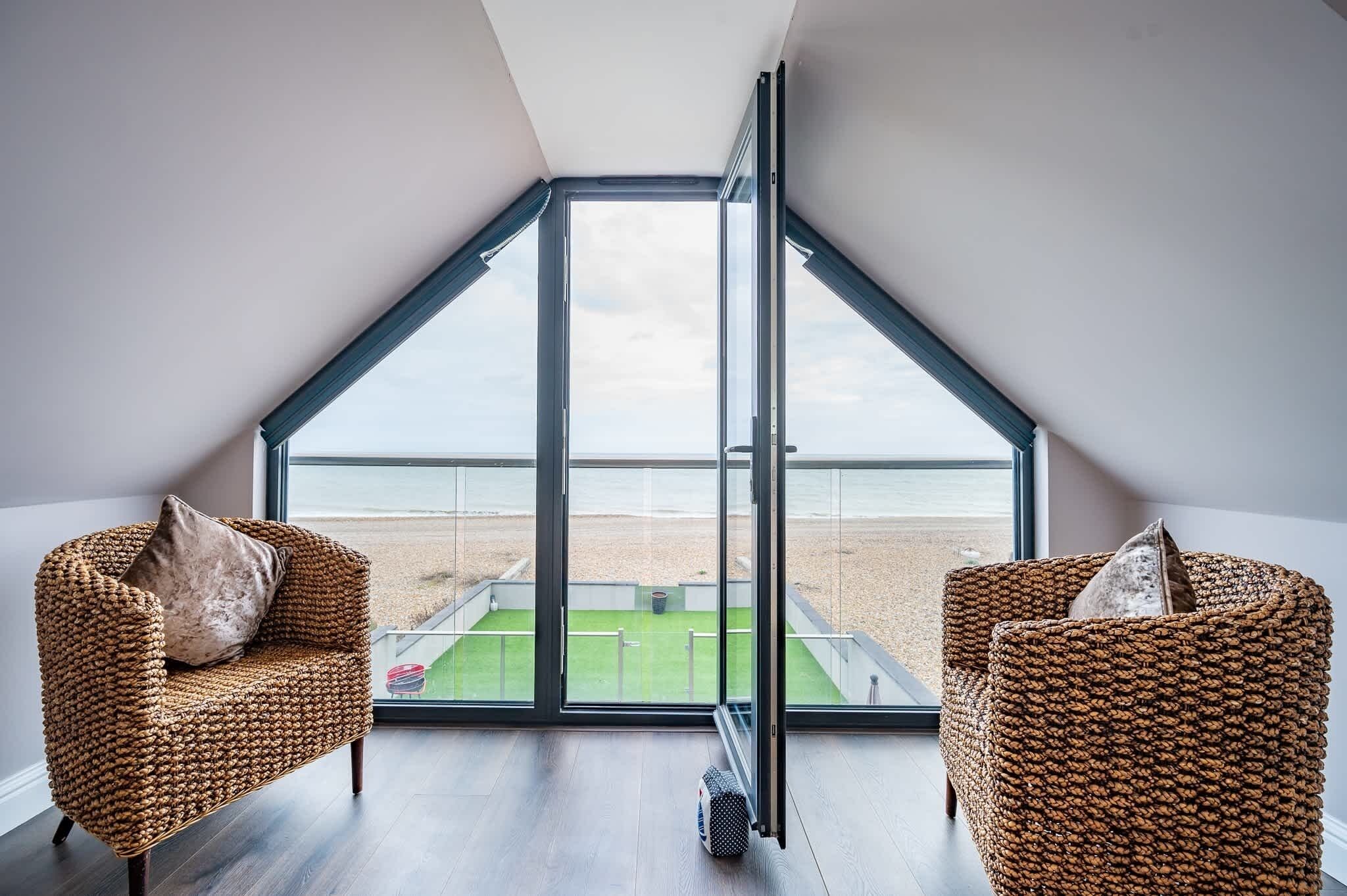 A room with a sloped ceiling, large glass windows, two wicker chairs, and a view of a beach.
