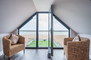 A room with a sloped ceiling, large glass windows, two wicker chairs, and a view of a beach.