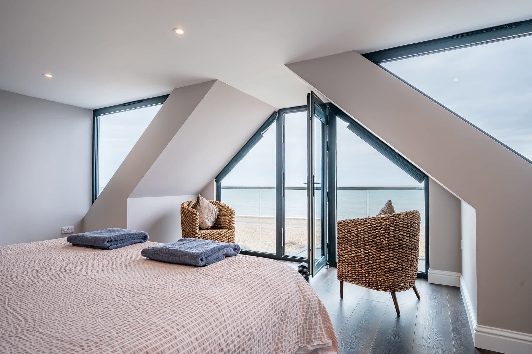 A bedroom with a large bed, two chairs, and a view of the sea through a large window.