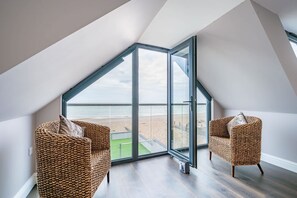 A room with a large glass window, two wicker chairs, and a view of the beach. - Oceanfront Villa with Stunning Beach Views (Pevensey)