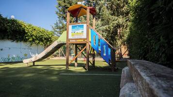 A playground with a slide and climbing structure, surrounded by greenery.
