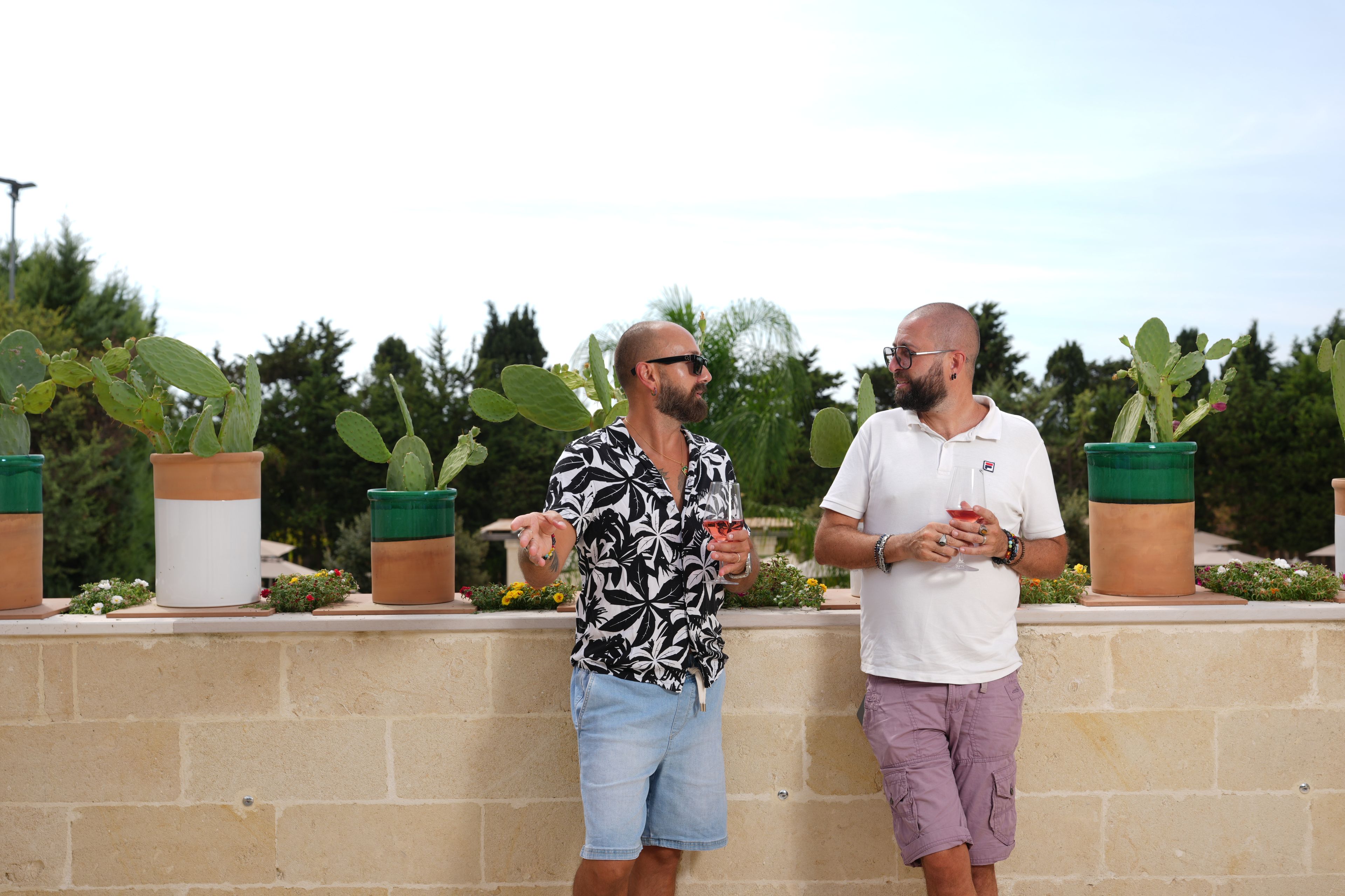 Two men standing outdoors, one in a patterned shirt and denim shorts, the other in a white shirt and purple shorts, holding drinks.