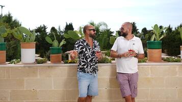 Two men standing outdoors, one in a patterned shirt and denim shorts, the other in a white shirt and purple shorts, holding drinks.