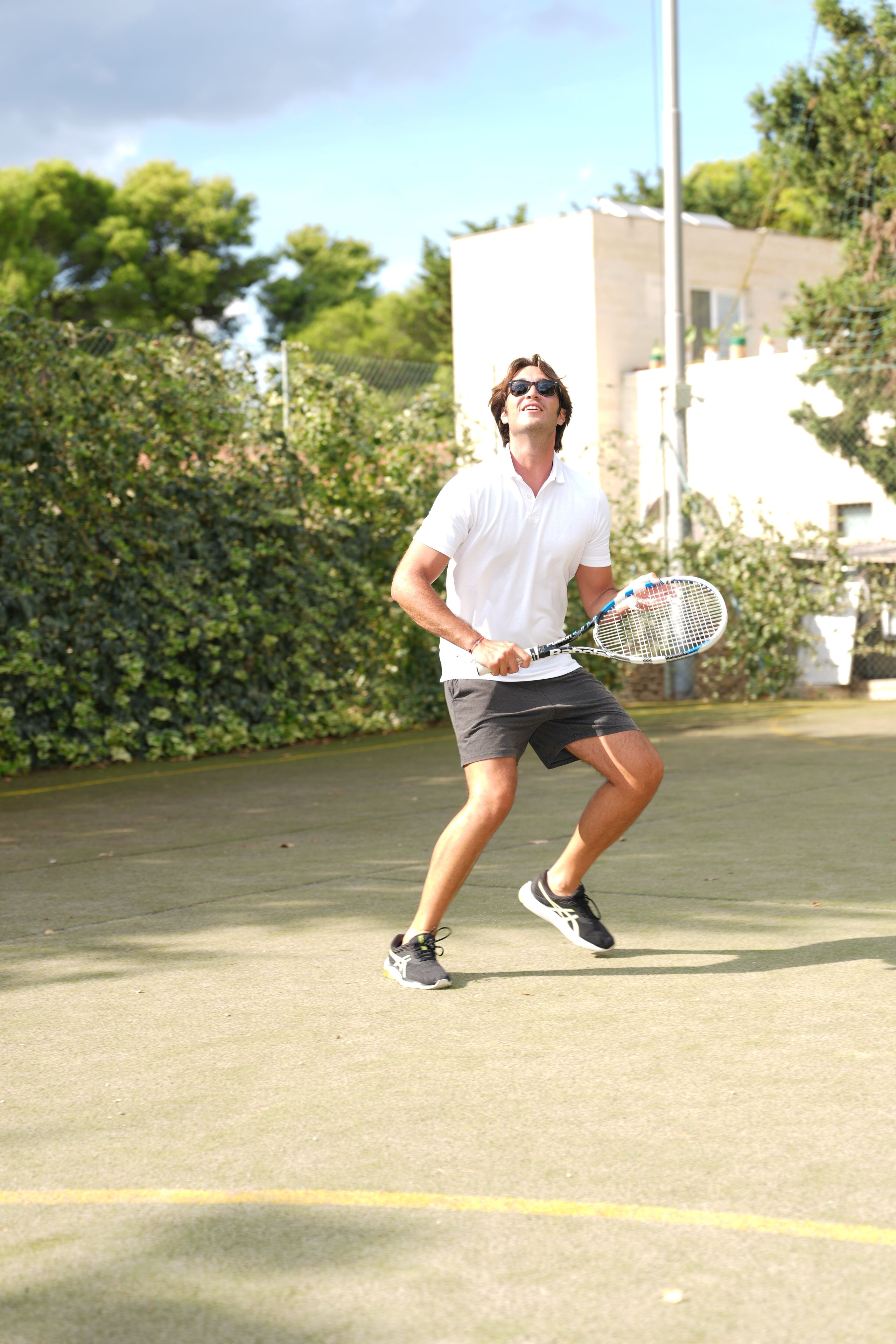 A person playing tennis on an outdoor court with a hedge and a building in the background.