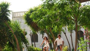 Two people riding bicycles on a paved path surrounded by lush greenery and a white building with arched windows.