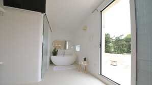 A modern bathroom with a freestanding bathtub, a small bedside table, and a large window offering a view of the outdoors. - Tenuta del Barco (Taranto)