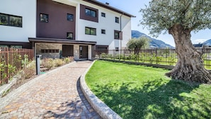 A modern multi-story building with a brick pathway leading to the entrance, surrounded by a well-maintained lawn and a large tree. - Apartment 'Sankt Martin' with Mountain View, Private Terrace and Wi-Fi (Sankt Martin)