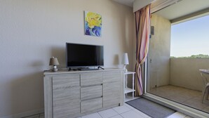 Una habitación con televisión sobre un mueble de madera, un cuadro en la pared y un balcón con una silla.