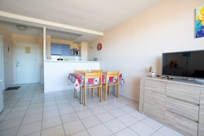 Uma sala de jantar com uma mesa posta para quatro pessoas, uma televisão sobre um móvel e uma cozinha ao fundo.