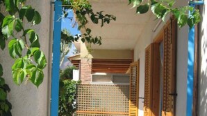 Zona exterior coberta amb una banca, una reixa i un edifici amb un mur arrebossat i persianes de fusta.