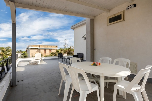 An outdoor patio with a round table and white plastic chairs, a covered area with a white ceiling, and a view of buildings in the background.