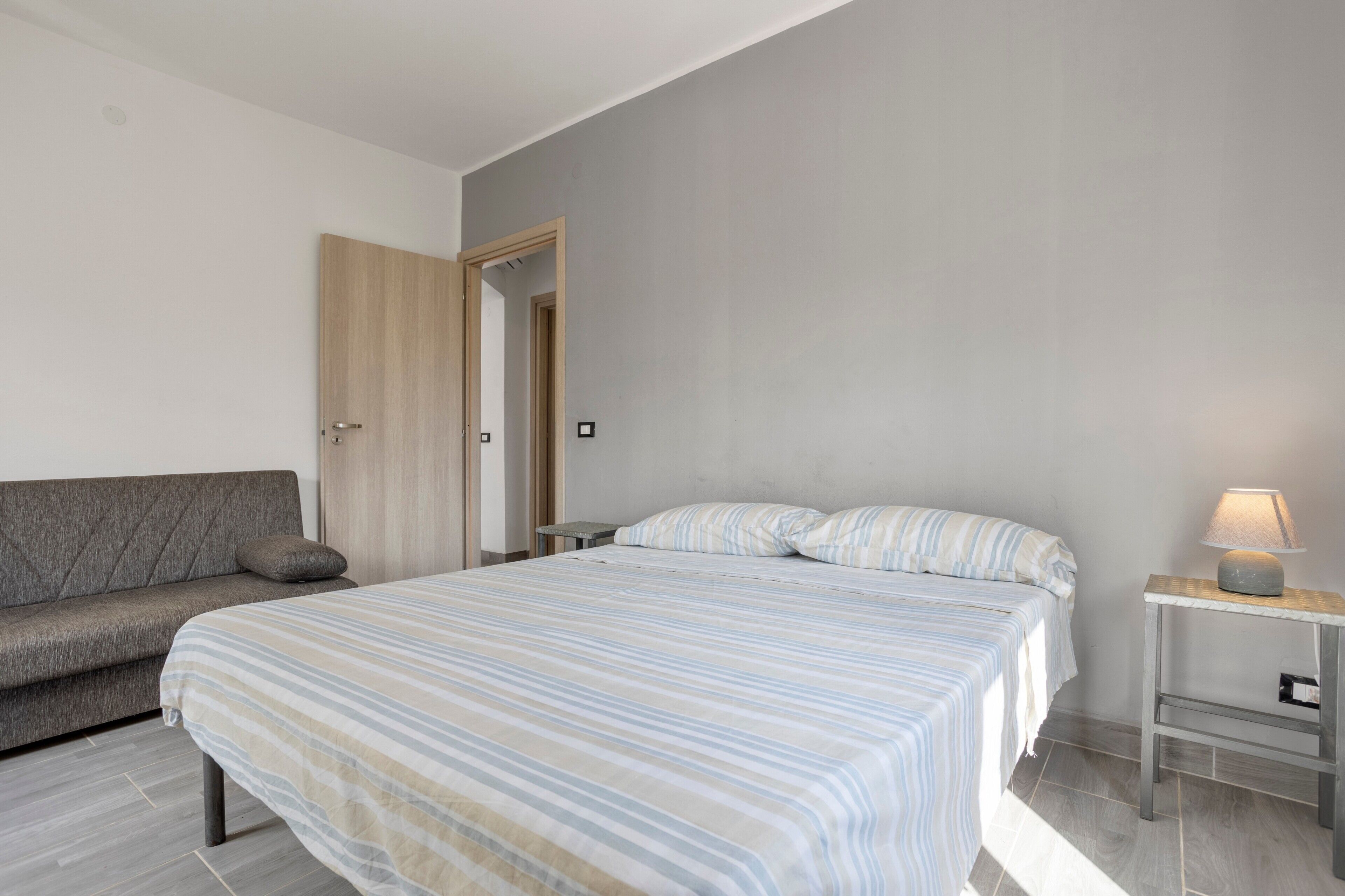 A bedroom with a bed, a bedside table with a lamp, a sofa, and a door.