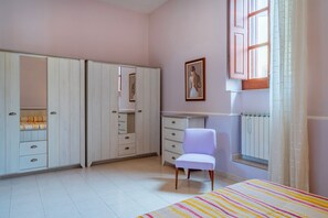 A bedroom with a bed, a chair, a wardrobe, a desk, a radiator, and a window with curtains.