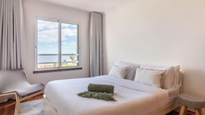 Un dormitorio con una cama grande, una silla, un taburete pequeño, una ventana con vistas y una cortina.
