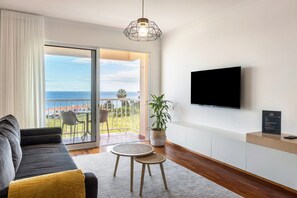 Un salón moderno con sofá, una mesita redonda, una planta en maceta, televisor y un balcón con vistas al océano.