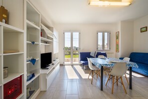 Een moderne woonkamer met een eethoek, inbouwplanken, een televisie en uitzicht door glazen deuren.