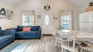 A living room with a blue sofa, a dining table with white chairs, a white door, and a clock above it.