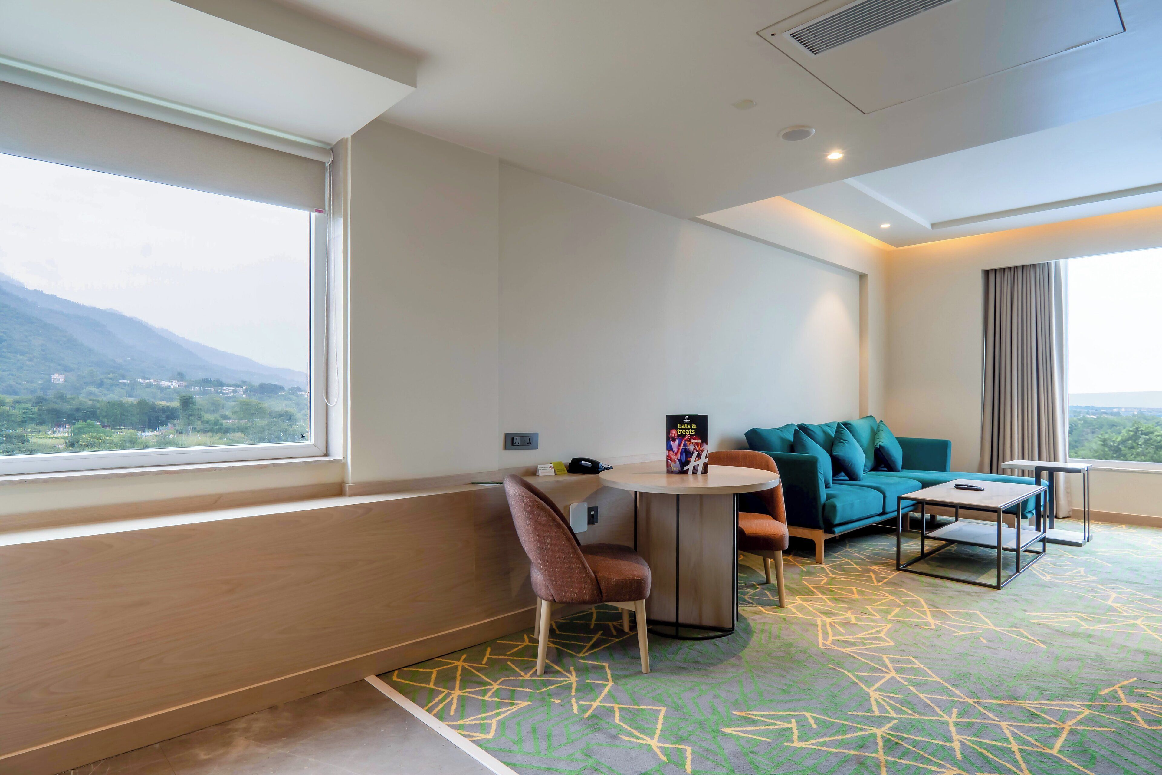 A modern hotel room with a large window, a round table, a sofa, and a patterned carpet.