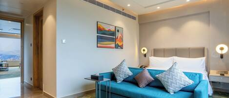 Quarto de hotel moderno com sofá verde-água, cama com roupa de cama branca, mesinha de cabeceira pequena e luminária fixada na parede.