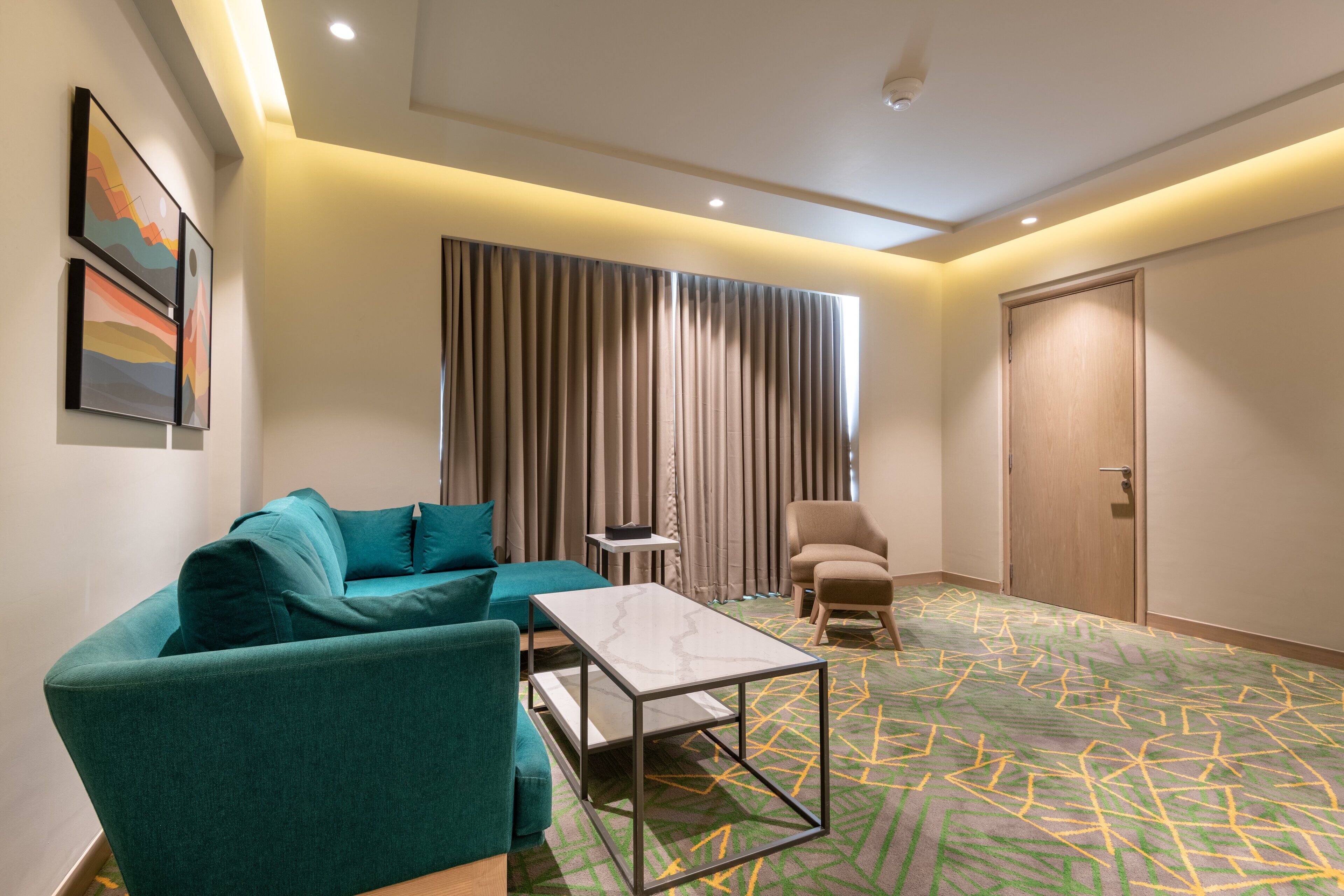 A modern hotel room with a sectional sofa, a small table, a chair, a large window with curtains, and a door.