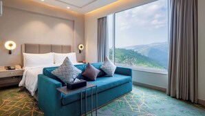 A hotel room with a bed, a blue sofa, a nightstand, and a large window with a view of mountains. - Holiday Inn Katra Vaishno Devi by IHG (Reasi)